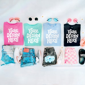 May include: Four blank t-shirts with the text "Your Design Here" in white on each shirt. The shirts are pink, light blue, turquoise, and black. The shirts are laid out on a white surface with various accessories, including sunglasses, denim shorts, checkered shorts, and a pair of black Vans shoes.