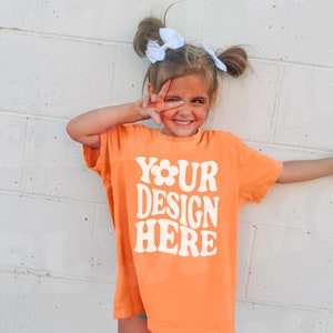 Puede incluir: Una niña pequeña con una camiseta naranja brillante con el texto "YOUR DESIGN HERE" en blanco. Está sonriendo y lleva el pelo en dos coletas con lazos blancos.
