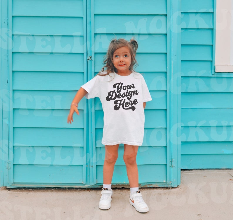 May include: A young person wearing a white t-shirt with the text "Your Design Here" in a retro font. The person is standing in front of a blue wooden wall.