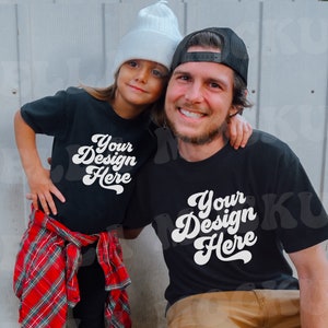 Pode incluir: Uma camiseta preta com o texto "Your Design Here" em uma fonte retrô. A camiseta é usada por uma pessoa e uma criança, ambos sorrindo. A criança usa um gorro branco e uma camisa xadrez vermelha amarrada na cintura.