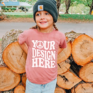 May include: A young person wearing a dusty rose t-shirt with the text "YOUR DESIGN HERE" in a white, retro font. The person is wearing a dark gray beanie and light blue jeans. The background includes a stack of cut logs.