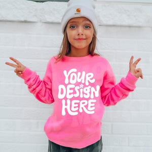 May include: A pink sweatshirt with the text "YOUR DESIGN HERE" in white. The sweatshirt has a crew neck and long sleeves. A child is wearing the sweatshirt and a white beanie.
