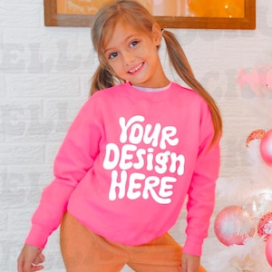 May include: A pink sweatshirt with the text "YOUR DESIGN HERE" in white. The sweatshirt is being worn by a young person with long brown hair.
