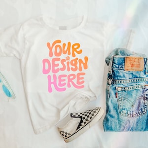 Könnte beinhalten: Weißes T-Shirt mit dem Text "YOUR DESIGN HERE" in rosa und orangefarbener Schrift. Das Shirt liegt auf einer weißen Oberfläche mit einer blauen Sonnenbrille, einem Paar schwarz-weiß karierten Sneakers und einer blauen Jeans.