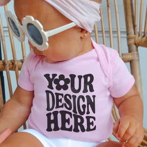 May include: A pink baby onesie with the text "YOUR DESIGN HERE" in black. The onesie is being worn by a baby wearing a white headband and white sunglasses.