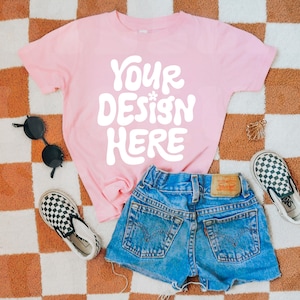 May include: A pink t-shirt with the text "YOUR DESIGN HERE" in white, laid out on a checkered rug with a pair of black sunglasses, a pair of black and white checkered slip-on shoes, and a pair of blue denim shorts.