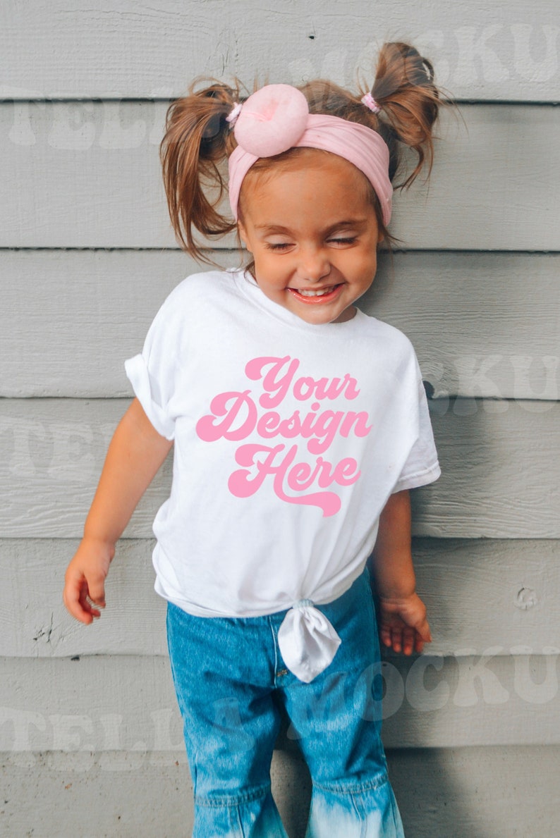 May include: A young child wearing a white t-shirt with the text "Your Design Here" in pink. The child is wearing blue jeans and a pink headband with a large pink pom-pom.