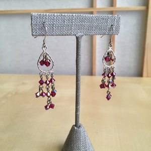 Puede incluir: Un par de pendientes de plata con cuentas de cristal rojo. Los pendientes están colgados de un soporte de tela.