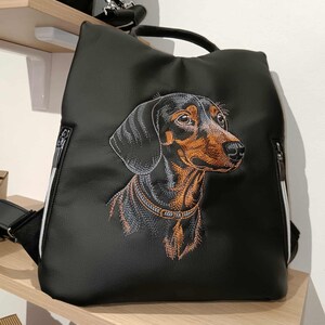Könnte beinhalten: Schwarzer Rucksack mit gesticktem Dackel-Design. Der Hund hat schwarzes und braunes Fell, detailliert mit Stickereien. Die Tasche hat einen oberen Griff, zwei seitliche Reißverschlüsse mit weißem Besatz und verstellbare Träger. Ein stilvolles Accessoire.