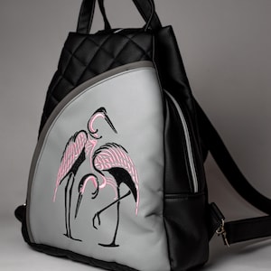 Könnte beinhalten: Schwarzer und grauer Rucksack mit einem gesteppten Oberteil und einer grauen Frontplatte. Die graue Platte zeigt ein gesticktes Design von drei rosa und schwarzen Flamingos. Der Rucksack hat einen oberen Griff und verstellbare Träger.