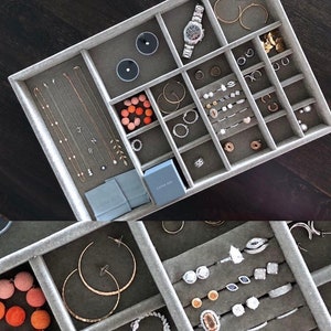 Puede incluir: Un organizador de joyas de terciopelo gris con múltiples compartimentos. El organizador está lleno de varias piezas de joyería, incluyendo collares, aretes, anillos y un reloj. El organizador también tiene una pequeña caja etiquetada como "Loma Boy".
