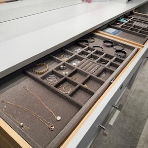 May include: A white wooden jewellery drawer organiser with a grey velvet lining, featuring multiple compartments for rings, necklaces, and bracelets. The tray contains various gold and silver-toned jewellery pieces, including chains, hoop earrings, and bangles.