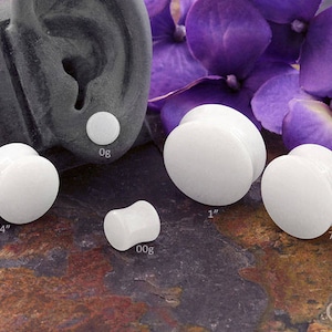 May include: A set of six white acrylic ear gauges in various sizes, including 0g, 00g, 3/4", 7/8", and 1". The gauges are displayed on a textured surface with purple flowers in the background.