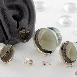 May include: A set of glass ear gauges in various sizes, ranging from 5/8 inch to 1 inch. The gauges are clear with a green iridescent sheen and have a black and white striped band around the edge. The gauges are shown on a white surface with a black ear model in the background.