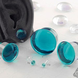 May include: A set of turquoise glass ear gauges in various sizes, including 5/8 inch, 0g, 1 inch, and 20mm. The gauges are displayed on a white surface with a black ear mold.