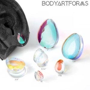 Può includere: Una collezione di tappi per le orecchie iridescenti a forma di goccia in varie misure, tra cui 2,5 cm, 2,2 cm, 1,9 cm, 1,4 cm, 0g e 2g. I tappi mostrano una gamma di colori e sono esposti su una superficie bianca. Il marchio "BODYARTFORMS" è visibile.