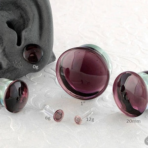 May include: A set of six glass ear gauges in various sizes, ranging from 9/16 inch to 20 mm. The gauges are made of clear glass with a purple hue and a smooth, polished finish.
