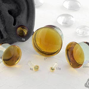 May include: Assortment of amber glass ear gauges in various sizes, including 0g, 5/8 inch, 8g, 12g, and 20mm. The gauges are displayed on a white surface with a black background.