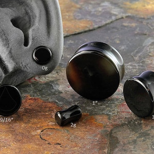 May include: A set of black obsidian ear gauges in various sizes, including 0g, 9/16", 1", 3/4", and 2g. The gauges are displayed on a textured stone surface.