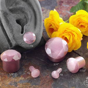 May include: A set of pink, faceted, gemstone plugs in various sizes, ranging from 0g to 7/16 inch. The plugs are displayed on a dark gray surface with yellow flowers.