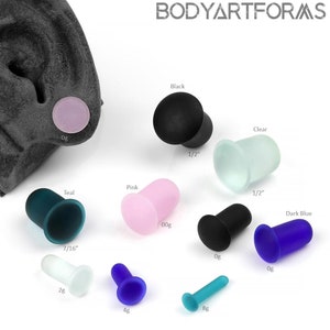 May include: A collection of colorful ear plugs in various sizes and colors, including black, clear, teal, pink, and dark blue. The plugs are labeled with sizes such as 0g, 00g, 2g, 4g, and 8g, and 1/2 inch and 7/16 inch. The brand name "BODYARTFORMS" is visible.