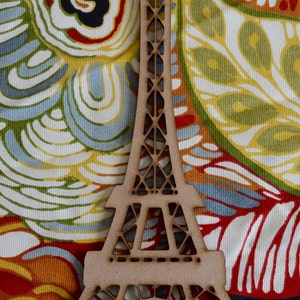 May include: A wooden cutout of the Eiffel Tower, painted in a light brown color, is placed on a colorful fabric with a paisley pattern.
