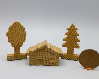 May include: A miniature wooden cabin with two trees and a penny. The cabin has a textured roof and a small door. One tree is a deciduous shape, the other is a pine tree shape. The penny is a copper color.