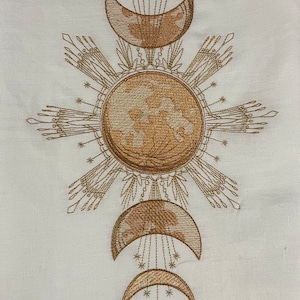 May include: Embroidered design featuring a sun with rays and four crescent moons in a symmetrical pattern. The design is in shades of brown on a white background.