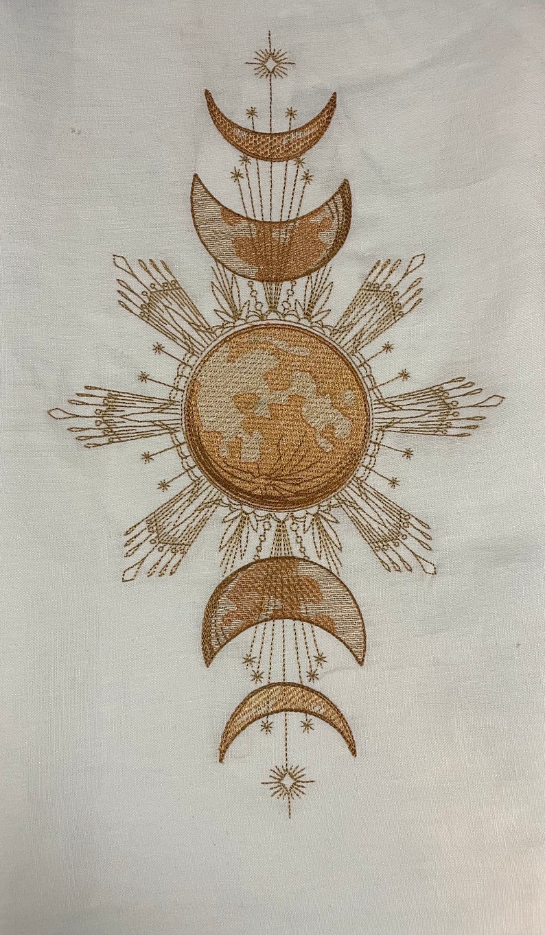 May include: Embroidered design featuring a sun with rays and four crescent moons in a symmetrical pattern. The design is in shades of brown on a white background.