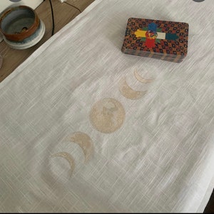 May include: A white linen altar cloth with a gold embroidered moon phases design. The cloth is laid flat on a surface and a deck of tarot cards is placed on top of the cloth.