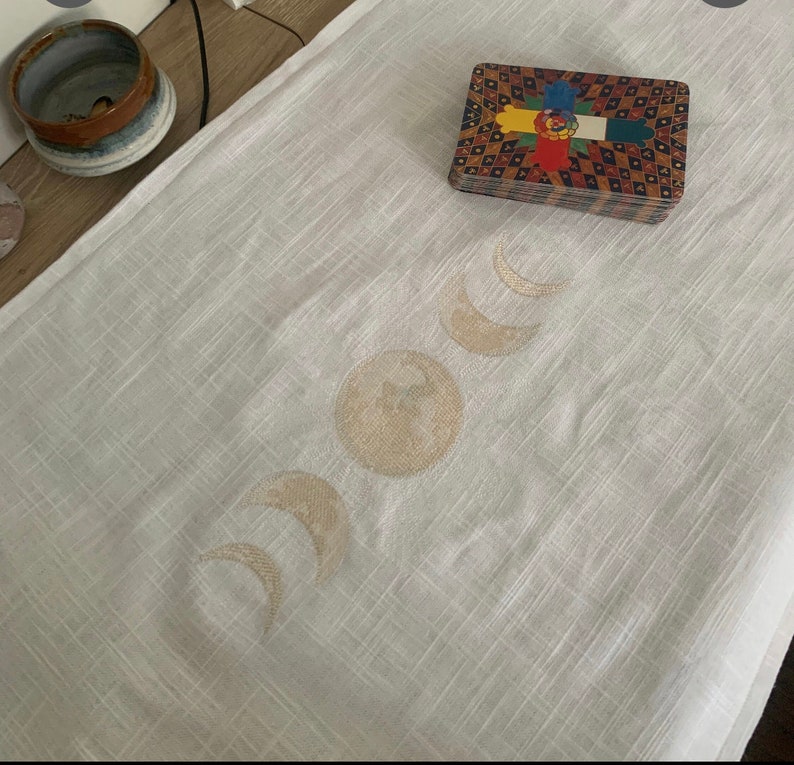 May include: A white linen altar cloth with a gold embroidered moon phases design. The cloth is laid flat on a surface and a deck of tarot cards is placed on top of the cloth.
