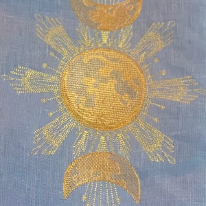 May include: A gold embroidered design on a light blue fabric featuring a sun with rays and a crescent moon with stars.