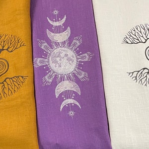 May include: Three linen tea towels with different designs. The first towel is mustard yellow with a black tree of life design. The second towel is purple with a white moon and sun design. The third towel is white with a black tree of life design.