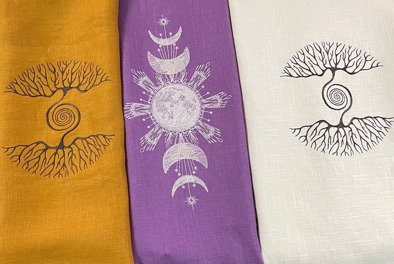 May include: Three linen tea towels with different designs. The first towel is mustard yellow with a black tree of life design. The second towel is purple with a white moon and sun design. The third towel is white with a black tree of life design.