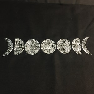 May include: A black fabric tote bag with a white moon phases design. The moon phases are in a crescent shape and are decorated with intricate details.