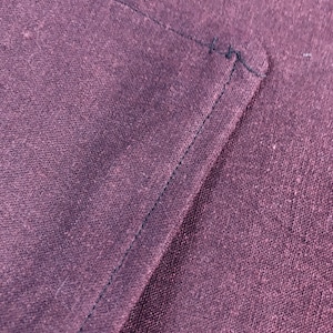 May include: Close-up of a dark purple fabric with a black stitched seam. The fabric has a textured, woven appearance.