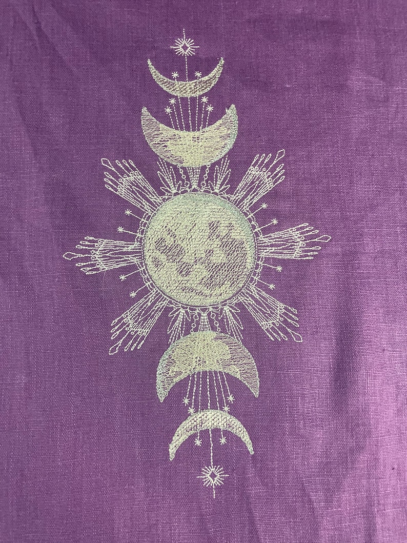 May include: A purple fabric with a white embroidered design of a sun and moon phases. The sun is surrounded by rays and the moon phases are arranged in a vertical line.