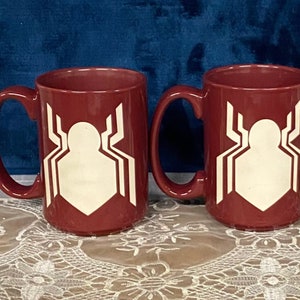 May include: Two maroon ceramic mugs with large white spider emblems. Each mug has a large handle and a cylindrical shape. The mugs are displayed on a white lace tablecloth against a blue background.