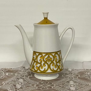 May include: White ceramic coffee pot with a gold and yellow ornate design around the base. The lid and top of the pot are gold. The pot has a long spout and a curved handle. The coffee pot is on a white lace tablecloth.