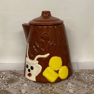 May include: Brown ceramic cookie jar shaped like a coffee pot with a white milk pitcher and yellow cookies on the front. The jar has the word "Cookies" written on the front.