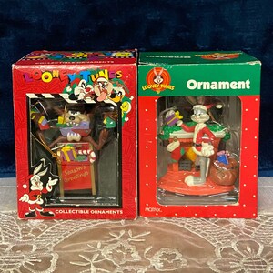 May include: Two Christmas ornament boxes featuring characters from the Looney Tunes cartoon. The box on the left features a red and green ornament of Tasmanian Devil holding a gift box. The box on the right features a red and green ornament of Bugs Bunny dressed as Santa Claus. Both boxes have the words "Looney Tunes" and "Collectible Ornaments" printed on them.
