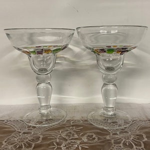 May include: Two clear glass margarita glasses with a colorful mosaic design around the rim. The glasses are on a white lace tablecloth.
