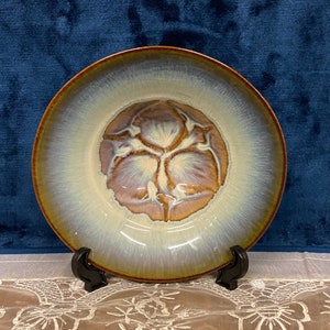 May include: A large, round ceramic plate with a light blue and brown glaze. The plate has a raised design in the center, resembling a stylized flower.