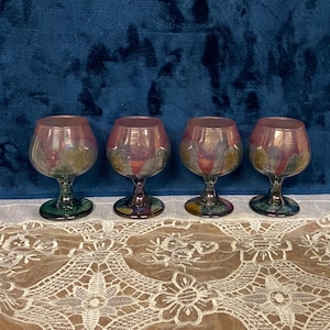 May include: Four iridescent glass goblets with a pink, green, and blue color scheme. The goblets are sitting on a white lace tablecloth.