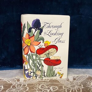 May include: A white ceramic book-shaped vase with a colorful illustration from "Through the Looking Glass." The design features mushrooms, flowers, and a sleeping cat. Text includes the title and author's name.