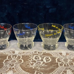 May include: Four clear glass tumblers with curved sides and a thick base. Each glass has text in a different color: red, blue, and yellow. The glasses are arranged on a white lace tablecloth against a dark blue background.