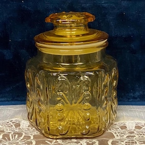 May include: A square amber glass canister with a matching lid. The glass features a raised, embossed scrollwork pattern on all four sides. The lid has a circular handle and a clear plastic seal around the rim to ensure a secure fit for the container.