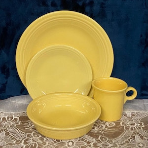May include: A set of yellow ceramic dinnerware, including a large plate, a smaller plate, a bowl, and a mug. The items are arranged on a white lace tablecloth against a dark blue background. The dinnerware has a classic, simple design.