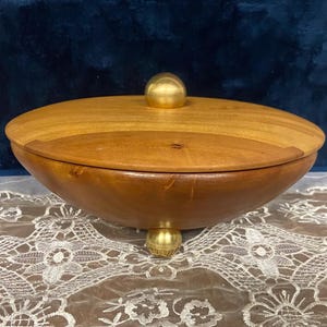 May include: A handcrafted wooden bowl with a lid. The bowl is a rich brown color with a lighter wood lid and a gold-colored knob. The bowl rests on three gold-colored feet. The bowl is sitting on a white lace tablecloth.