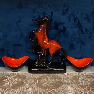 May include: A ceramic horse sculpture with brown and orange glaze, standing on a black base. Two small orange and black ceramic bowls are on either side of the sculpture.